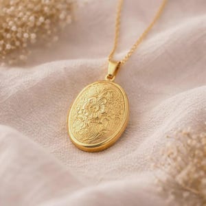 Peut inclure: Un pendentif médaillon ovale doré suspendu à une fine chaîne assortie. Le médaillon présente un motif floral en relief complexe sur sa face avant, mettant en valeur des pétales détaillés et des motifs de vignes entrelacées.