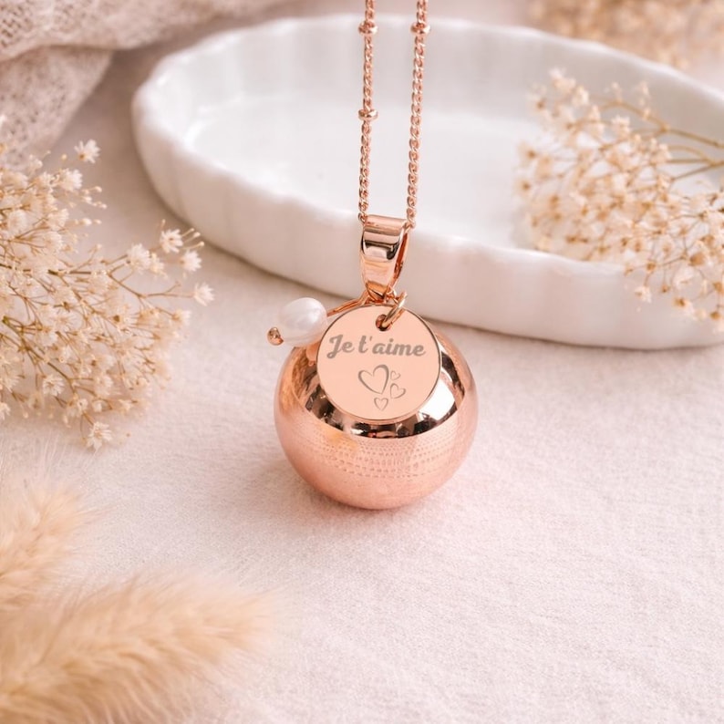 May include: Rose gold pendant necklace featuring a spherical charm with a textured surface. A small circular charm is engraved with the words "Je t'aime" and two hearts. A pearl bead accents the design. The necklace hangs from a delicate chain.