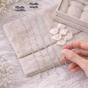 May include: A collection of silver necklaces with different chain styles, including a satellite chain and a simple chain. The necklaces are displayed on a neutral-colored fabric, with the words "Maille satellite" and "Maille simple" visible.