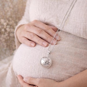 Peut inclure: Un collier pendentif argenté avec un charm rond et poli. Le collier est placé sur un ventre de femme enceinte, suggérant un thème de maternité ou de bébé. La chaîne est délicate et le charm reflète la lumière.