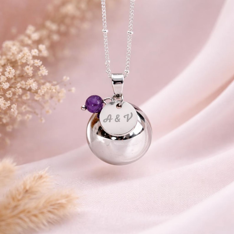 May include: A silver pendant necklace featuring a large, polished sphere with a smaller disc engraved with "A & V". A small, purple gemstone is attached. The necklace hangs on a delicate chain.