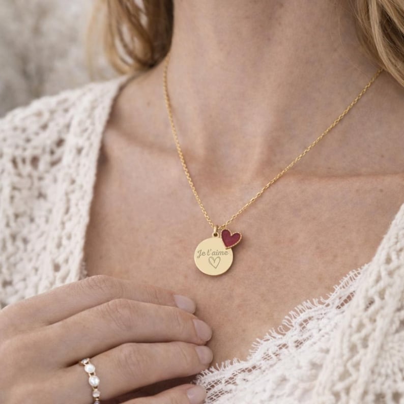 May include: A gold-colored necklace with two pendants. One pendant is a small, round disc with the words "Je t'aime" and a heart symbol. The other pendant is a red heart. The necklace is worn around the neck.