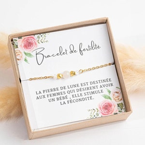 Op de afbeelding: Een gouden kettingarmband met witte en gouden kralen in een klein doosje met een bloemenpatroon en de tekst "Bracelet de fertilité" en "LA PIERRE DE LUNE EST DESTINÉE AUX FEMMES QUI DÉSIRENT AVOIR UN BÉBÉ, ELLE STIMULE LA FÉCONDITÉ."
