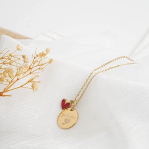 May include: A gold-colored necklace with a chain and two pendants. One pendant is a small red heart, and the other is an oval with the engraved phrase "Je t'aime" and two heart symbols. The necklace is displayed on a white fabric.