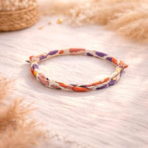 Puede incluir: Una pulsera de tela con un estampado floral en tonos naranja, morado y rosa. La pulsera es circular y parece estar hecha de tela trenzada. El fondo es una tela blanca suave.