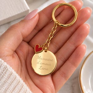 Może przedstawiać: Złoty metalowy brelok do kluczy z okrągłą zawieszką wygrawerowaną napisem Marraine d'amour Lana. Brelok zawiera małą czerwoną zawieszkę w kształcie serca z koralikową obwódką przymocowaną do złotego łańcuszka oraz okrągłe kółko na klucze.