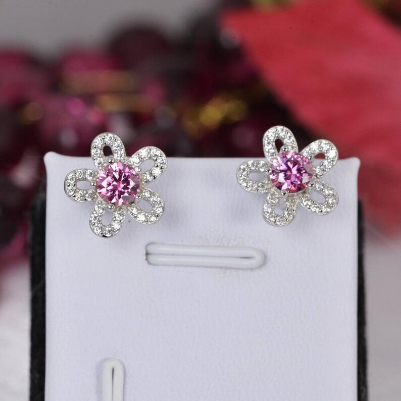 May include: A pair of flower-shaped stud earrings. Each earring features a pink gemstone center surrounded by a halo of small, clear stones. The earrings are set in a silver-colored metal and are displayed on a white surface.