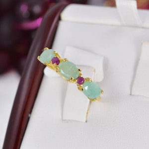 May include: A gold ring featuring three oval-shaped light green stones alternating with two small round dark pink stones. The stones are set in a row along the thin gold band, creating a colorful and symmetrical design on the jewelry piece.