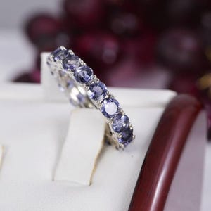 May include: A silver-toned eternity band featuring a continuous row of round, faceted, light purple-blue gemstones set in a prong setting. The ring is displayed at an angle, showcasing the uniform size and circular cut of each individual stone.