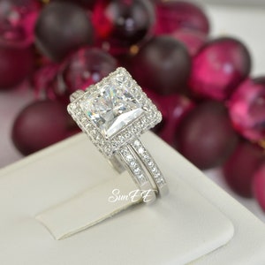 May include: A silver engagement ring set with a large square-cut cubic zirconia stone surrounded by smaller stones. The ring is on a white display stand.