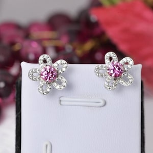 May include: A pair of flower-shaped stud earrings. Each earring features a pink gemstone center surrounded by small, clear gemstones. The earrings are set in a silver-colored metal and are displayed on a white surface.