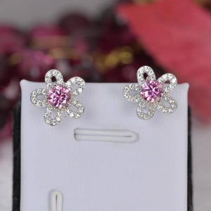 May include: A pair of flower-shaped stud earrings. Each earring features a pink gemstone center surrounded by small, clear gemstones, creating a floral design. The earrings are set on a white background, showcasing their delicate details.