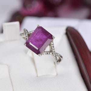 May include: A silver ring featuring a large, rectangular, deep magenta-colored gemstone set in the center. The band is designed with a crisscross pattern and is encrusted with small, clear, sparkling stones along the shoulders of the ring.
