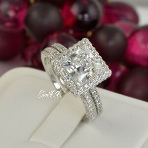 May include: A silver engagement ring set with a large, square-cut diamond surrounded by smaller diamonds. The ring is on a white display stand.