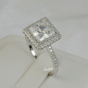 May include: A silver ring with a large square-shaped clear cubic zirconia stone surrounded by a double halo of smaller clear cubic zirconia stones. The band is also set with smaller clear cubic zirconia stones.