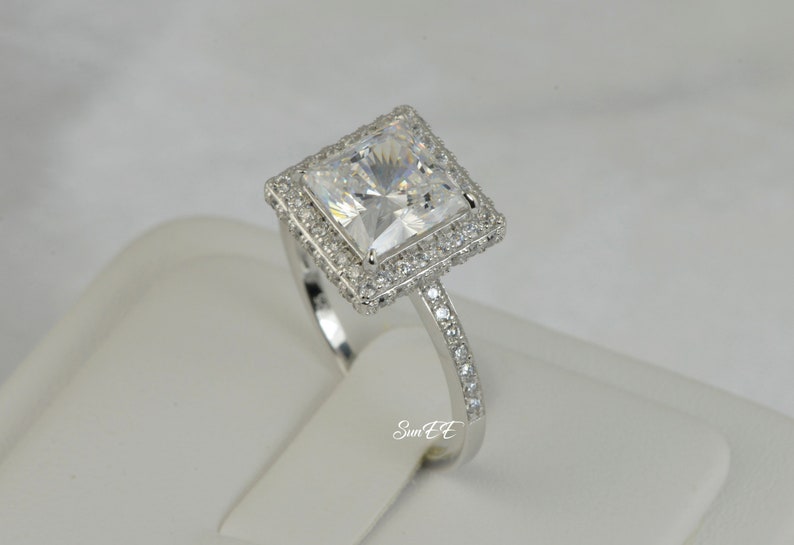 May include: A silver ring with a large square-shaped clear cubic zirconia stone surrounded by a double halo of smaller clear cubic zirconia stones. The band is also set with smaller clear cubic zirconia stones.