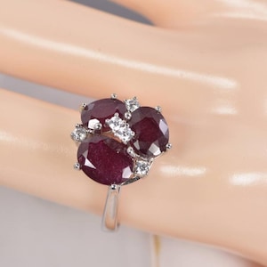 May include: A silver-toned ring featuring three oval-cut deep red rubies arranged in a cluster, accented with small, clear, round-cut white stones set between the rubies on a polished metal band.