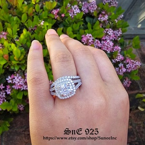 Puede incluir: Un anillo de plata con una piedra grande, redonda y transparente rodeada de piedras más pequeñas. El anillo tiene un diseño de triple banda con una banda retorcida en la parte inferior.