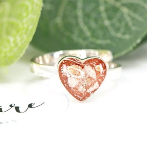 May include: A silver ring with a heart-shaped red and white resin inlay. The resin contains small white flecks and is set in a silver heart-shaped bezel.