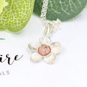 Peut inclure: Un collier avec un pendentif en forme de fleur argentée avec une pierre rose au centre. La fleur a cinq pétales et une surface texturée.