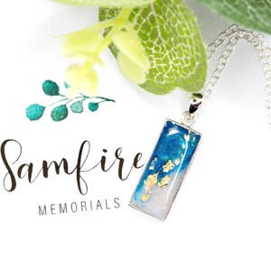 Peut inclure: Un collier en chaîne argent avec un pendentif rectangulaire en résine bleue et blanche avec des paillettes dorées. Le pendentif est sur un fond blanc avec le texte "Samfire MEMORIALS".