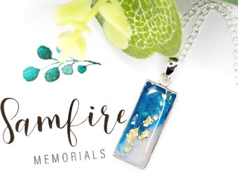 Può includere: Una collana con catena d'argento con un ciondolo rettangolare in resina blu e bianca con scaglie dorate. Il ciondolo è su uno sfondo bianco con il testo "Samfire MEMORIALS".