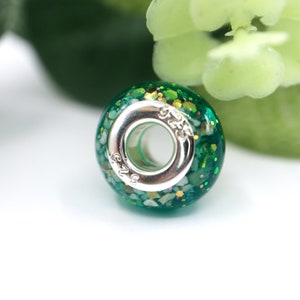 May include: A green glass bead with silver accents and a sparkly finish. The bead has a hole in the center for threading onto a bracelet or necklace. The bead is marked with the number "925" on the side.