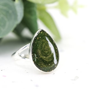 May include: A silver ring with a teardrop-shaped green stone. The stone has gold glitter embedded in it.