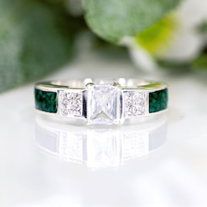 May include: A silver ring featuring a large, rectangular, clear gemstone at its centre, with two smaller square gemstones either side. The band has dark green inlays. The ring is shown on a reflective surface. The ring's design is elegant.