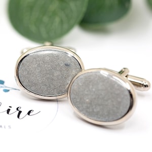 May include: A pair of oval-shaped cufflinks with a silver metal frame and a gray concrete-like inlay.