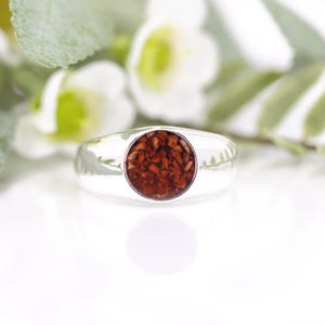 Könnte beinhalten: Ein silberner Ring mit einem kreisförmigen, rotbraunen Steineinsatz. Der Ring ist vor einem verschwommenen Hintergrund aus weißen Blüten und grünen Blättern platziert, was eine natürliche und elegante Ästhetik erzeugt. Das Design des Rings ist schlicht, aber auffällig.