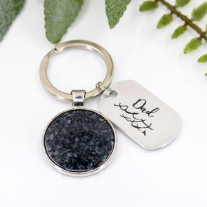 Puede incluir: Llavero plateado con un colgante redondo moteado azul oscuro y gris y una etiqueta rectangular. La etiqueta está grabada con la palabra "Dad" en cursiva y un pequeño diseño. Un regalo sentimental.