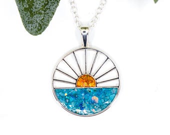 Può includere: Collana con pendente color argento con un design circolare raffigurante un'alba sul mare. La metà superiore del pendente ha raggi bianchi e un sole arancione, la metà inferiore è blu con glitter. Un gioiello delicato.