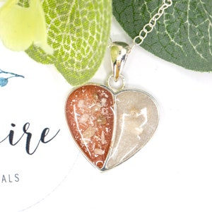 May include: A silver heart-shaped pendant necklace with two halves filled with different colored sand. One half is filled with white sand and the other half is filled with pink and brown sand.