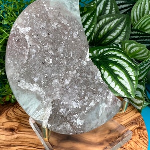 May include: A large, half-moon shaped amethyst geode with a clear, crystalline interior. The geode is displayed on a clear acrylic stand with a gold metal base.