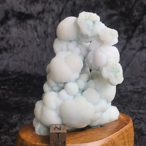 Pode incluir: Um espécime mineral branco e azul, com bolhas, sobre uma base de madeira. Um cubo de 1 cm está incluído para escala.