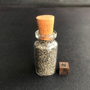 May include: A small glass bottle with a cork stopper filled with black sand. A 1 cm cube is next to the bottle for scale.
