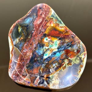 May include: A polished, irregularly shaped stone with a vibrant display of colors. The stone features a mix of red, blue, yellow, and green hues, with intricate patterns and textures. The surface is smooth and reflective, showcasing the natural beauty of the mineral.