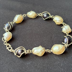 Puede incluir: Pulsera hecha a mano con perlas crema y gris oscuro, cada una engastada en alambre dorado. La pulsera está unida con pequeños eslabones de cadena dorados, creando un diseño delicado. Las perlas miden aproximadamente 0,8 cm de diámetro.