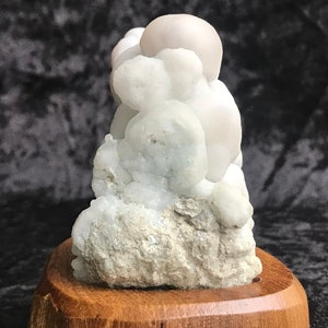 Puede incluir: Un espécimen mineral blanco y burbujeante sobre una base de madera. El espécimen tiene un grupo de formaciones redondas y blancas.
