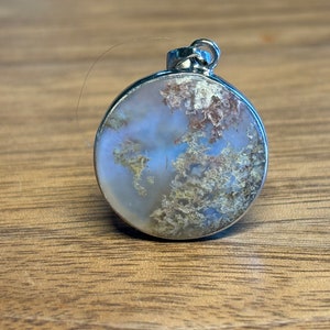 May include: Round pendant with a silver-colored metal frame. The pendant features a light blue and white background with brown and green moss-like inclusions. The pendant has a loop at the top for a chain.