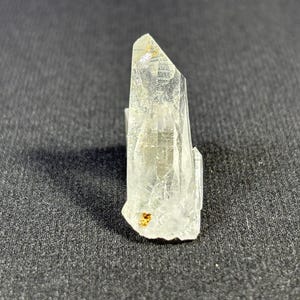 May include: A clear quartz crystal with a pointed tip and a rough, textured surface. The crystal is about 2 inches tall and has a slightly yellow hue.