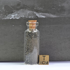 Puede incluir: Un pequeño frasco de vidrio lleno de arena negra, con un tapón de corcho. Una regla de 1 cm está al lado del frasco.