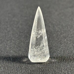 Può includere: Una punta di cristallo di quarzo trasparente con una punta appuntita e una superficie liscia e lucida. Il cristallo ha un aspetto nuvoloso e lattiginoso con inclusiones visibili.