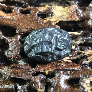 以下が含まれることがあります： 複雑な彫刻が施された黒い楕円形の石。石は、茶色と黒のまだら模様の表面を持つ木の板の上に置かれています。