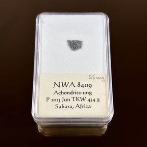 Puede incluir: Un pequeño fragmento de meteorito achondrita-ung de color gris oscuro dentro de una vitrina de plástico transparente. La vitrina tiene un interior blanco y una etiqueta beige con texto negro que dice "NWA 8409 Achondrite-ung Sahara, Africa". El peso es de 55mg.