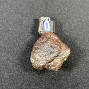 May include: A small, brown, irregularly shaped rock with a silver metal loop attached to the top. The loop has a small, white number "0" on it.