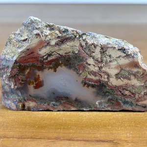 Könnte beinhalten: Ein natürlich geformtes Stein-Exemplar mit einer Vielzahl von Farben. Der Stein weist einen zentralen, hellfarbigen Bereich auf, der von Bändern in Rot-, Braun- und Grautönen umgeben ist. Die Oberfläche ist strukturiert und rau.