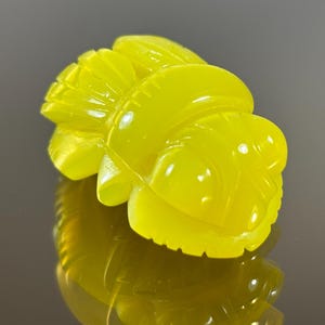 May include: A carved, yellow gemstone shaped like a stylized bee. The gemstone has a glossy finish and is highly reflective, with intricate details carved into its surface. The bee is positioned at an angle, showcasing its form.