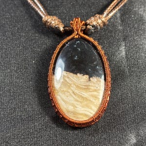 Pode incluir: Colar com pingente oval com uma pedra preta e bege, envolta em fio de cobre. O pingente pende de um cordão castanho. A pedra tem um padrão único. Uma joia marcante.
